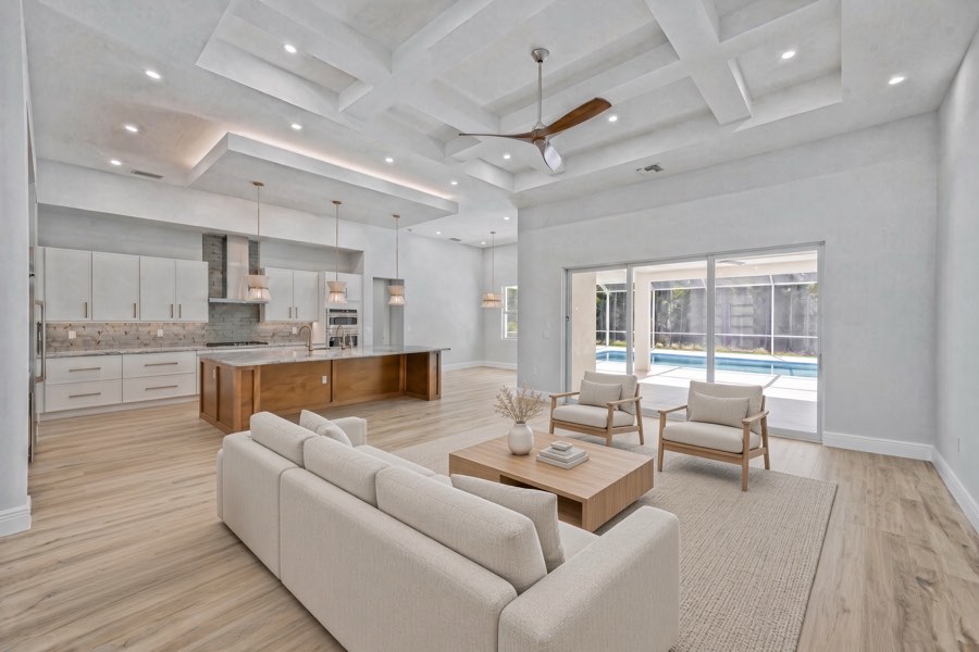 Sala amplia y luminosa con sofá blanco, techo artesonado, isla de cocina y puertas corredizas hacia la piscina